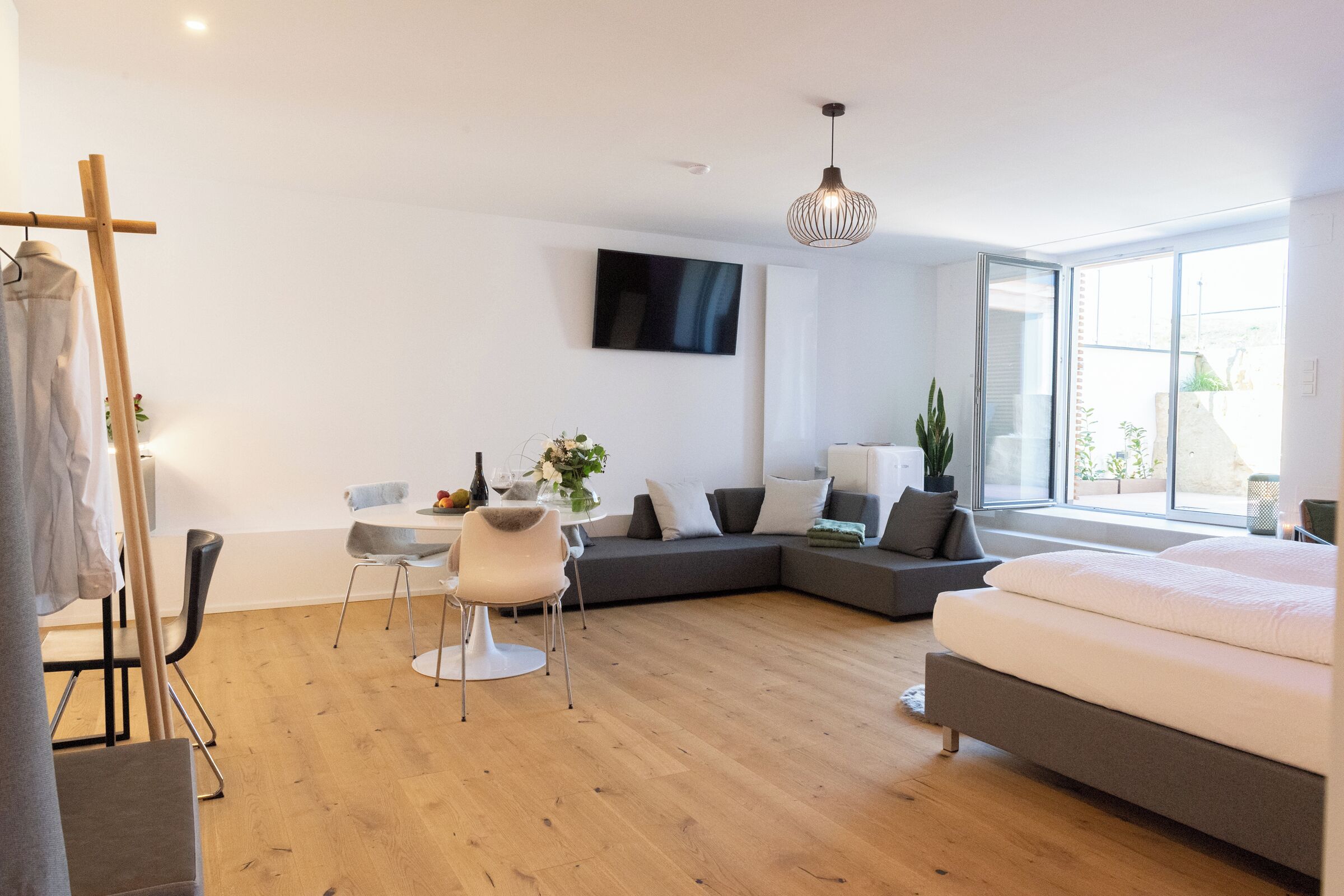 A modern, well-lit room with a large bed, a sofa, a dining table with chairs, a TV mounted on the wall, and a large window providing natural light.