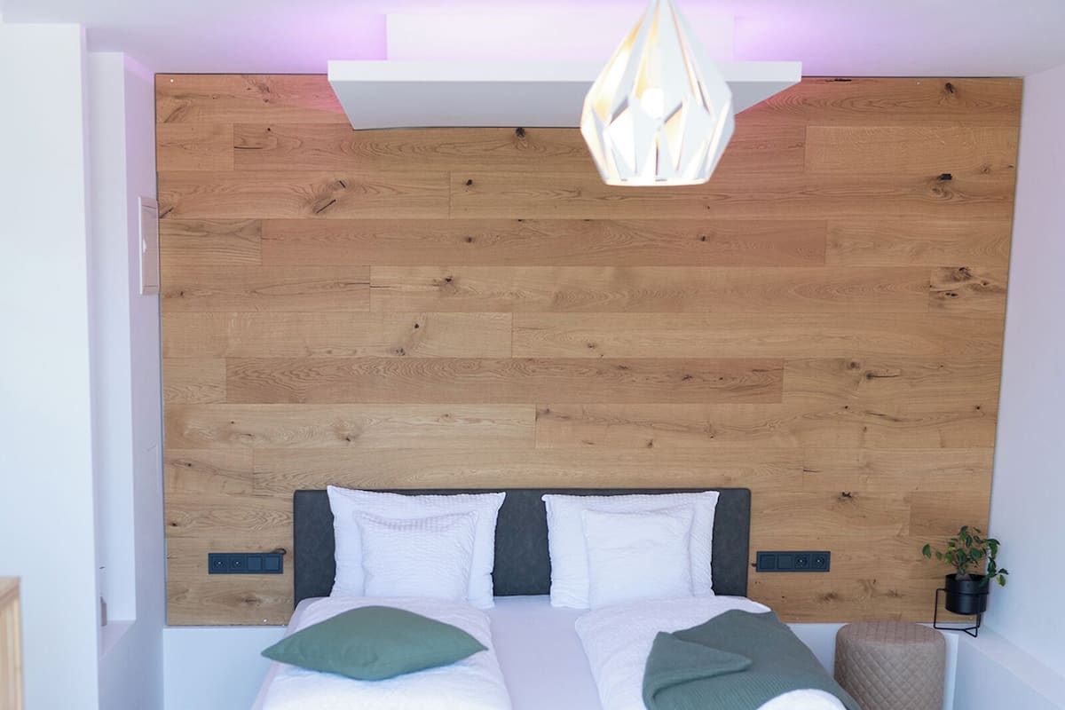 Un dormitorio moderno con cabecera de madera, cama con ropa de cama blanca, un banco gris y una lámpara colgante.