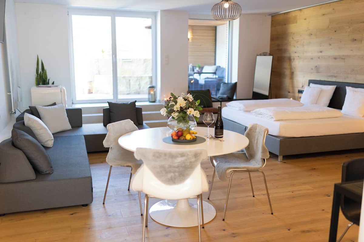 Una habitación de hotel moderna con un sofá, una mesa de comedor con frutero, una cama y vistas al exterior.