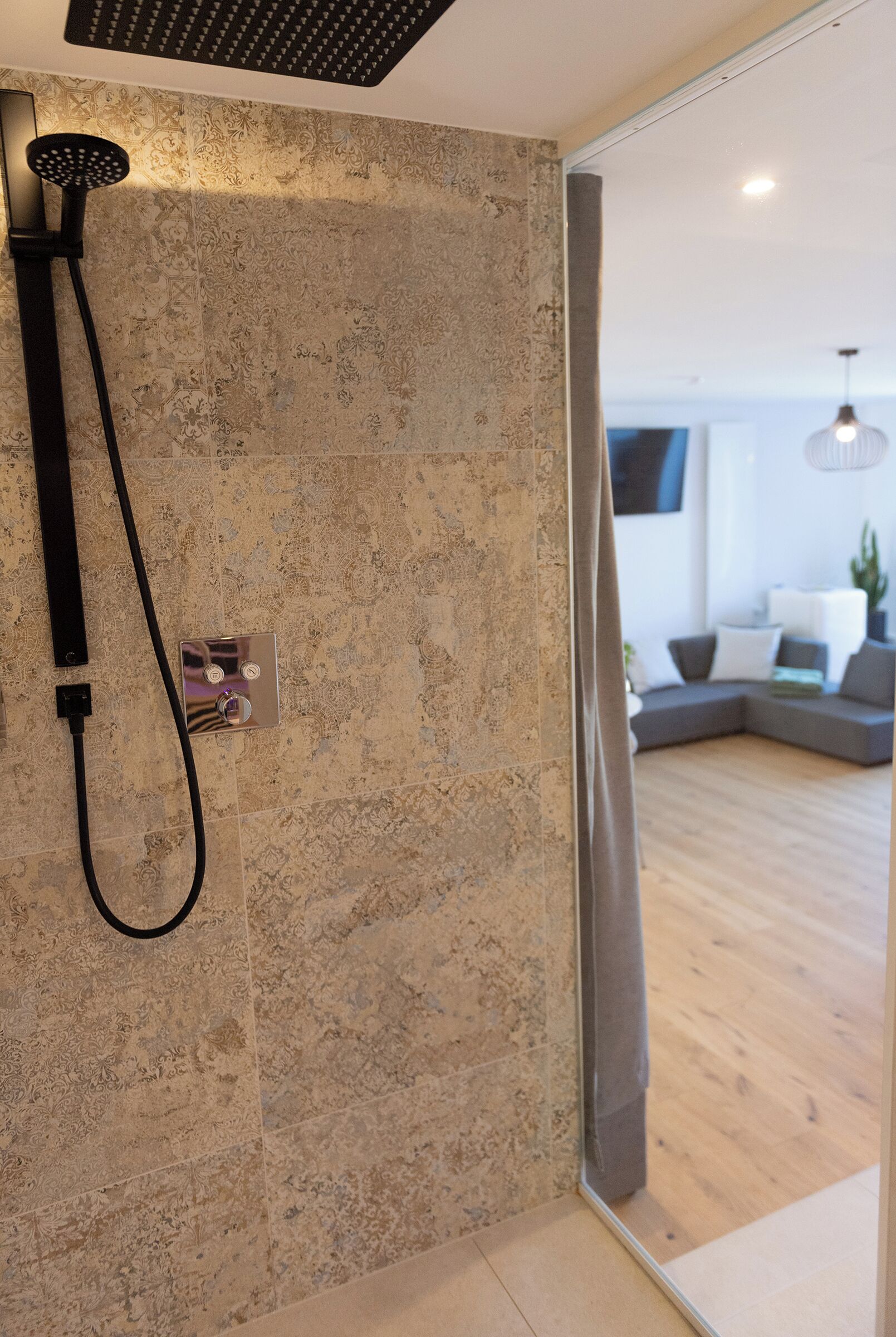 A modern bathroom with a shower, a control panel, and a glass door leading to a living area with a sofa and a plant.