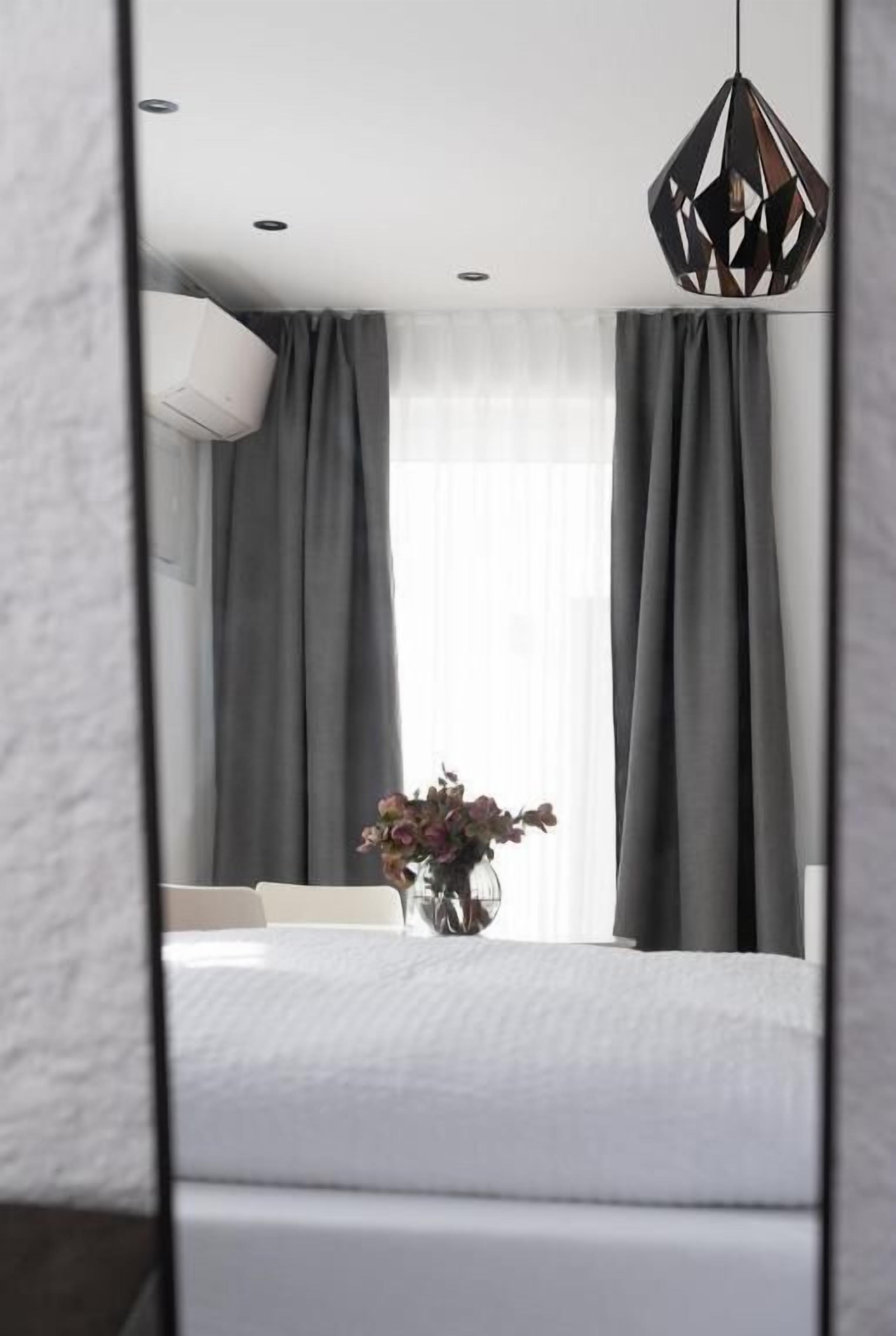 A modern bedroom with a large bed, grey curtains, a hanging light fixture, and a vase with flowers on the windowsill.