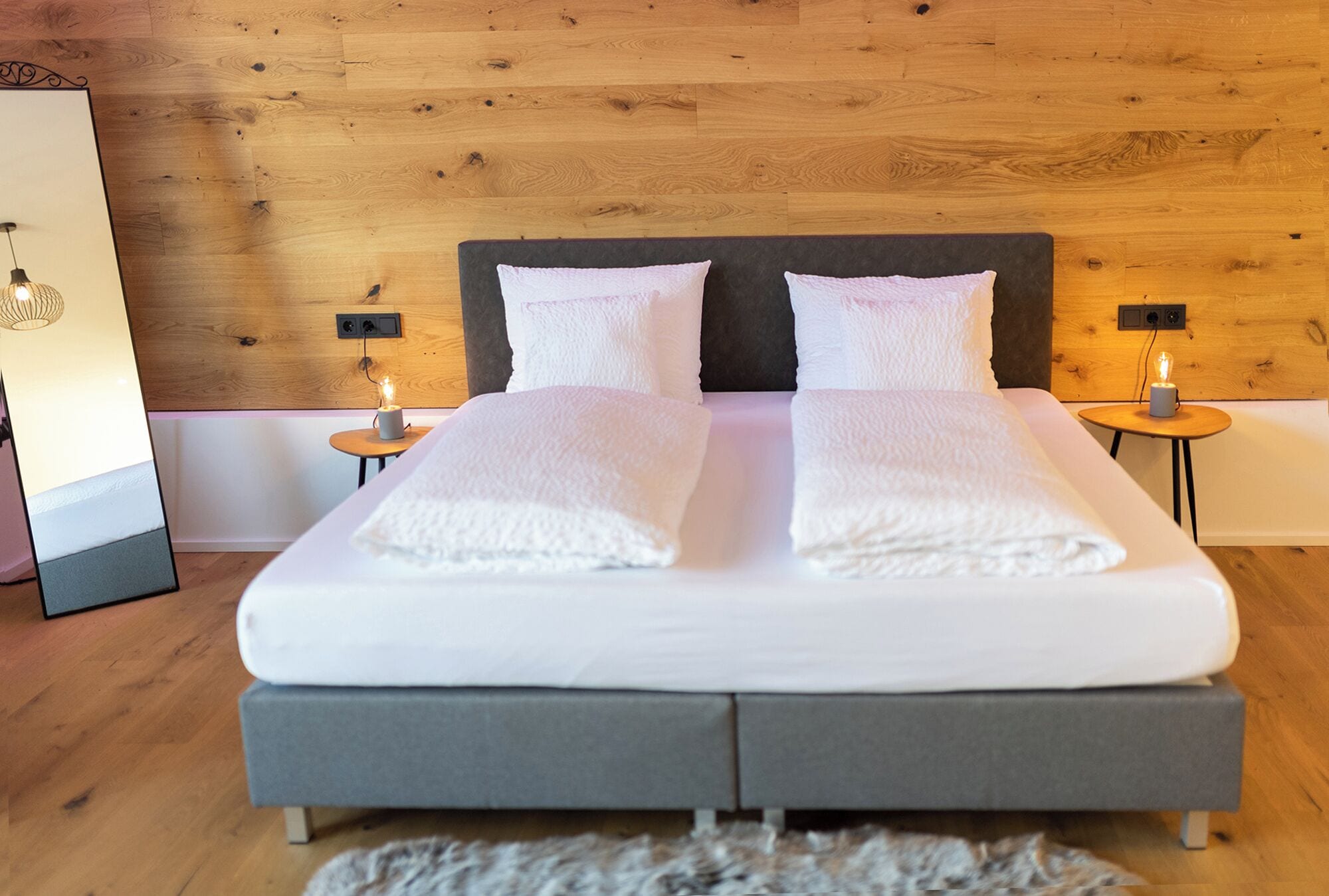 Een net opgemaakt bed met witte lakens, twee nachtkastjes met kaarsen en houten lambrisering.