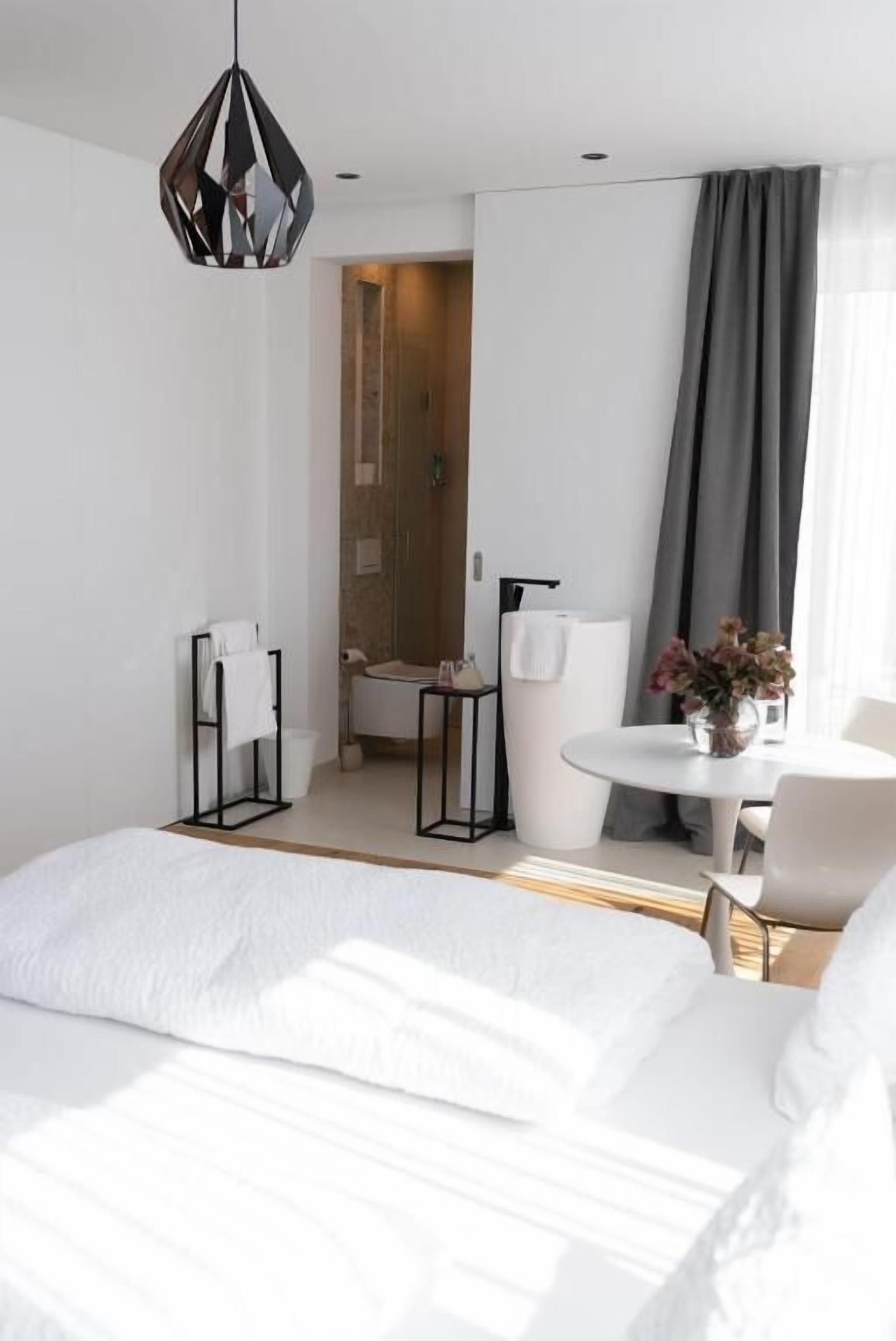 A modern hotel room with a large bed, a round dining table, a small white chair, a hanging pendant light, and a view of the bathroom through an open door.