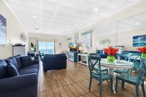 A spacious living area with a sofa, a dining table with chairs, and a view of the ocean through large windows. - 🌊🌴Gulf Views | Walk to Beach + Peg Leg Pete's | Sans Souci 207 (Pensacola Beach)