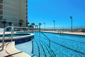 A swimming pool with a hot tub, surrounded by palm trees and a fence, with a tall building in the background. - 🌊🌴Gulf Views | Walk to Beach + Peg Leg Pete's | Sans Souci 207 (Pensacola Beach)