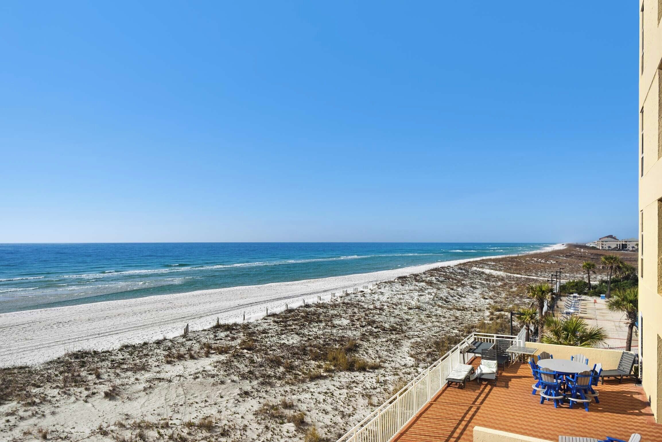 A beach view from a balcony with a wooden deck, outdoor seating, and a clear view of the ocean.