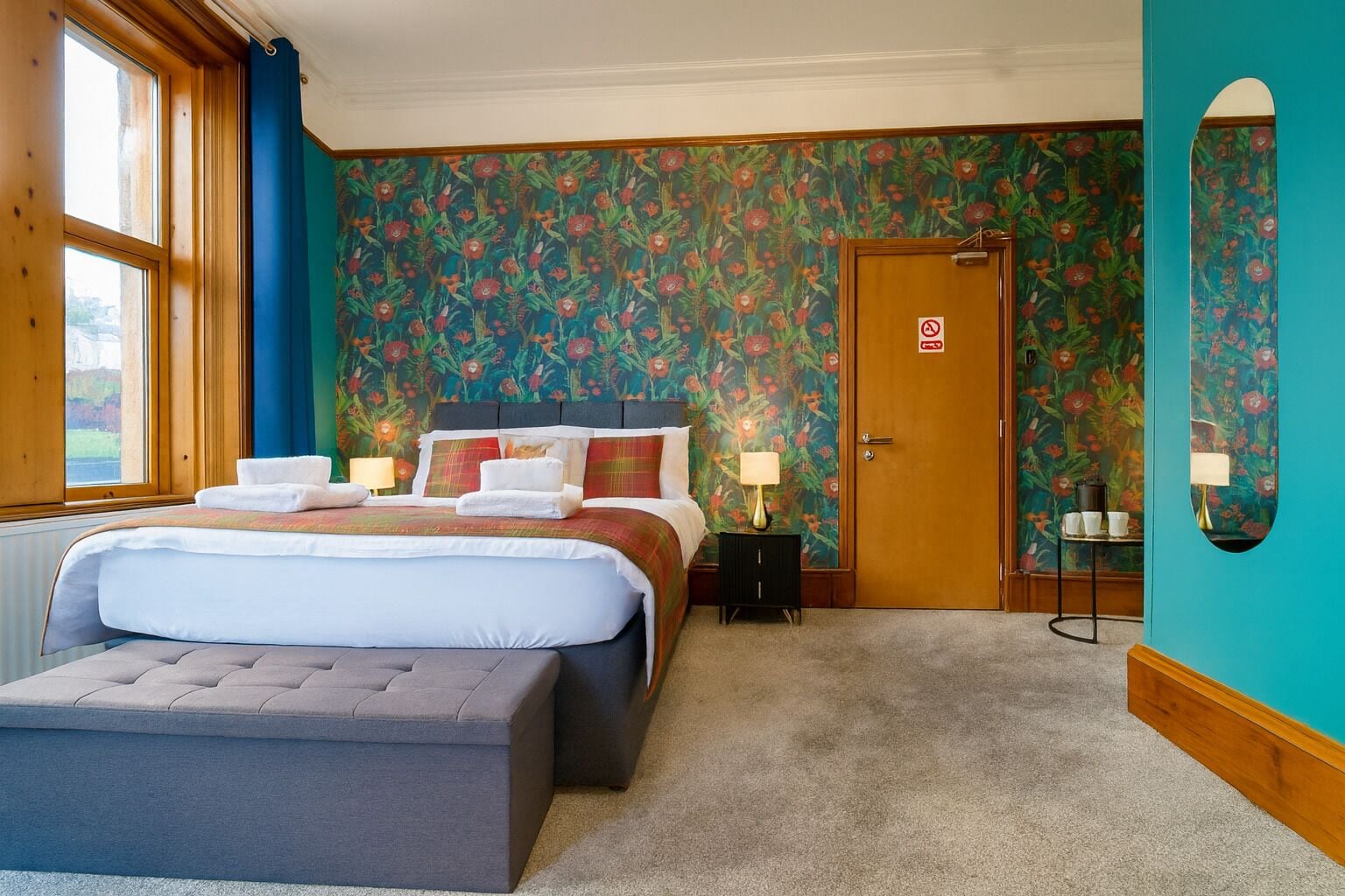 A bedroom with a large bed, a bedside table with a lamp, a door with a no smoking sign, and a window with curtains.