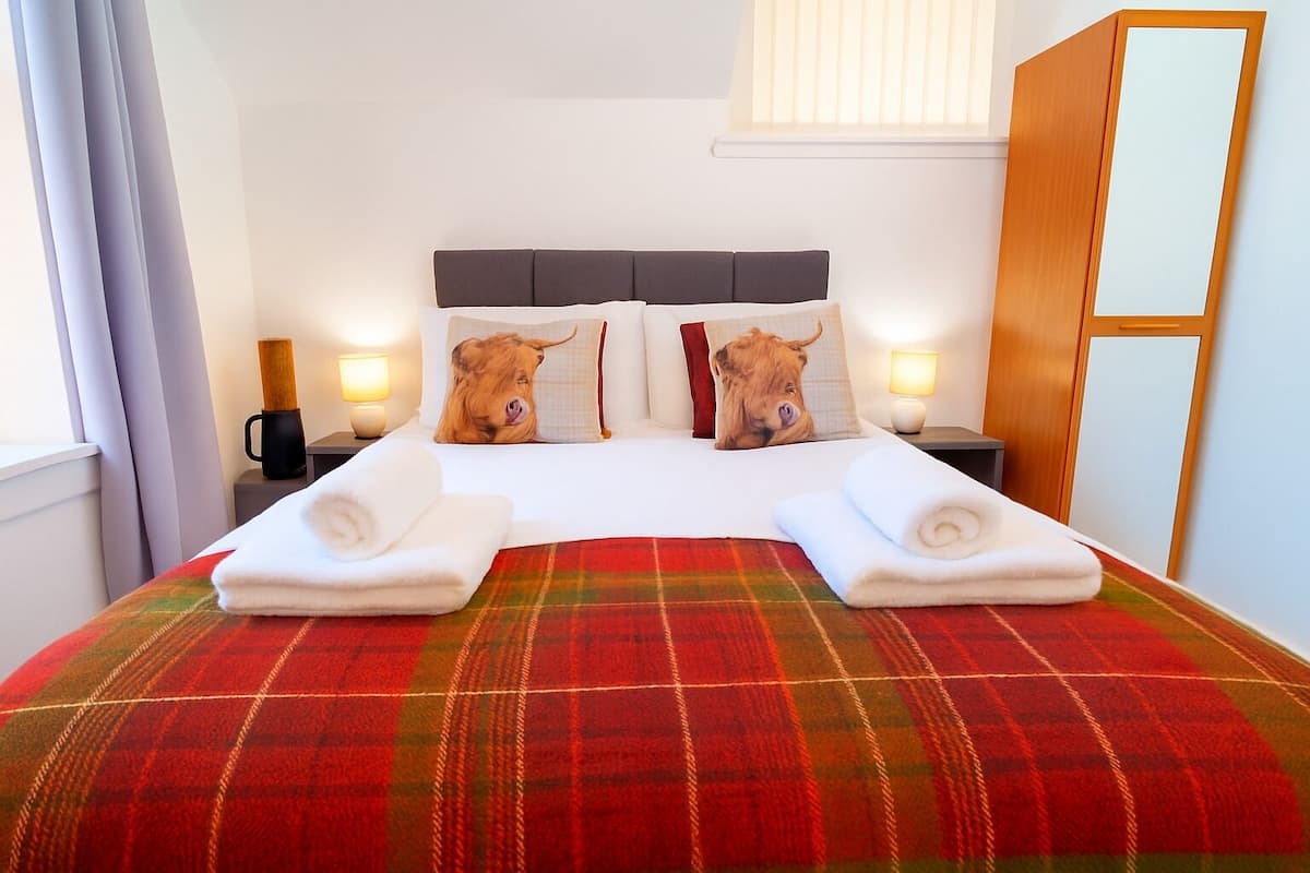 Habitación de hotel con cama, dos almohadas con diseños de animales, manta escocesa, dos toallas enrolladas, lámparas de noche y un armario de madera.