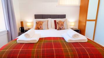 Hotelkamer met een bed, twee kussens met dierenprints, een geruit plaid, twee opgerolde handdoeken, bedlampen en een houten garderobe.