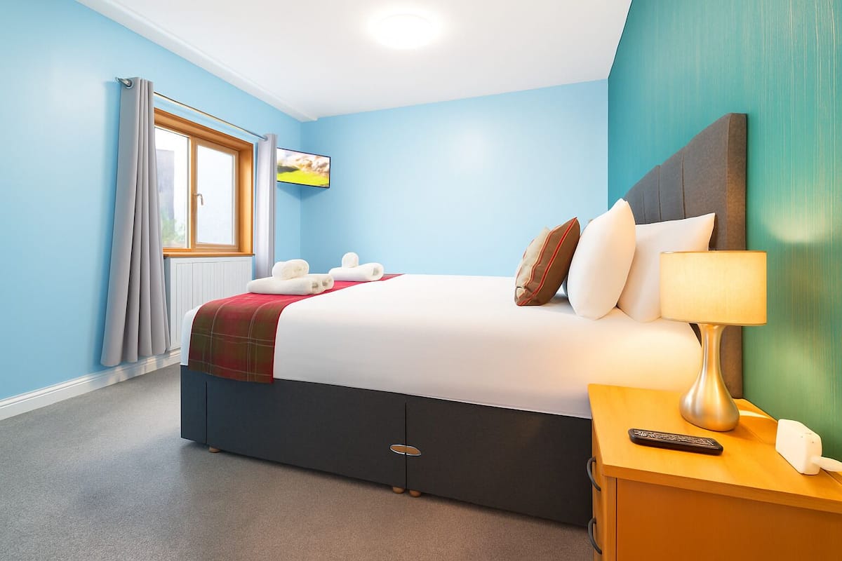Una habitación de hotel con cama, mesita de noche con lámpara, control remoto y ventana con cortinas.