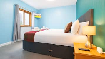 Quarto de hotel com uma cama, mesa de cabeceira com abajur, controle remoto e uma janela com cortinas.