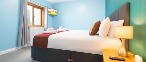 A hotel room with a bed, a nightstand with a lamp, a remote control, and a window with curtains.