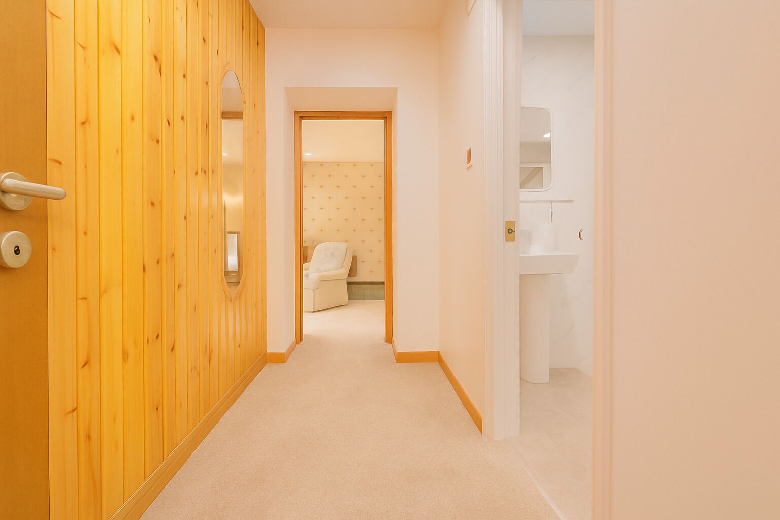 A hallway with a wooden door, a white wall with a cross pattern, and a bathroom with a sink and mirror.