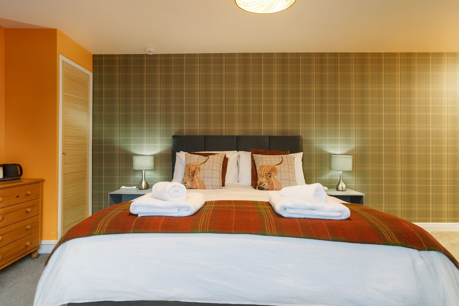 A hotel room with a bed, two nightstands, a lamp, a dresser, and a plaid-patterned wall.
