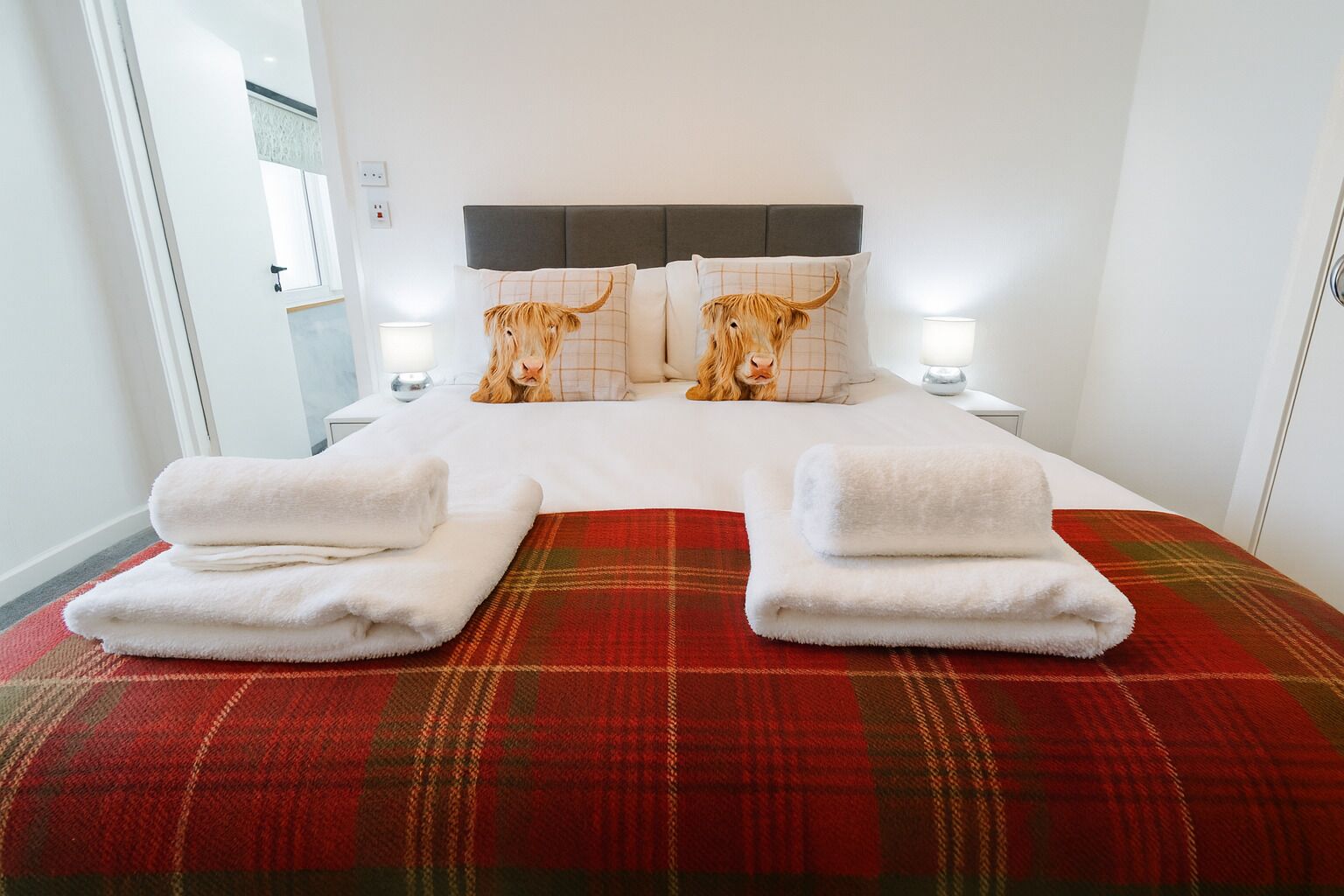 Habitación de hotel con cama, dos mesitas de noche, colcha roja de cuadros y dos toallas.