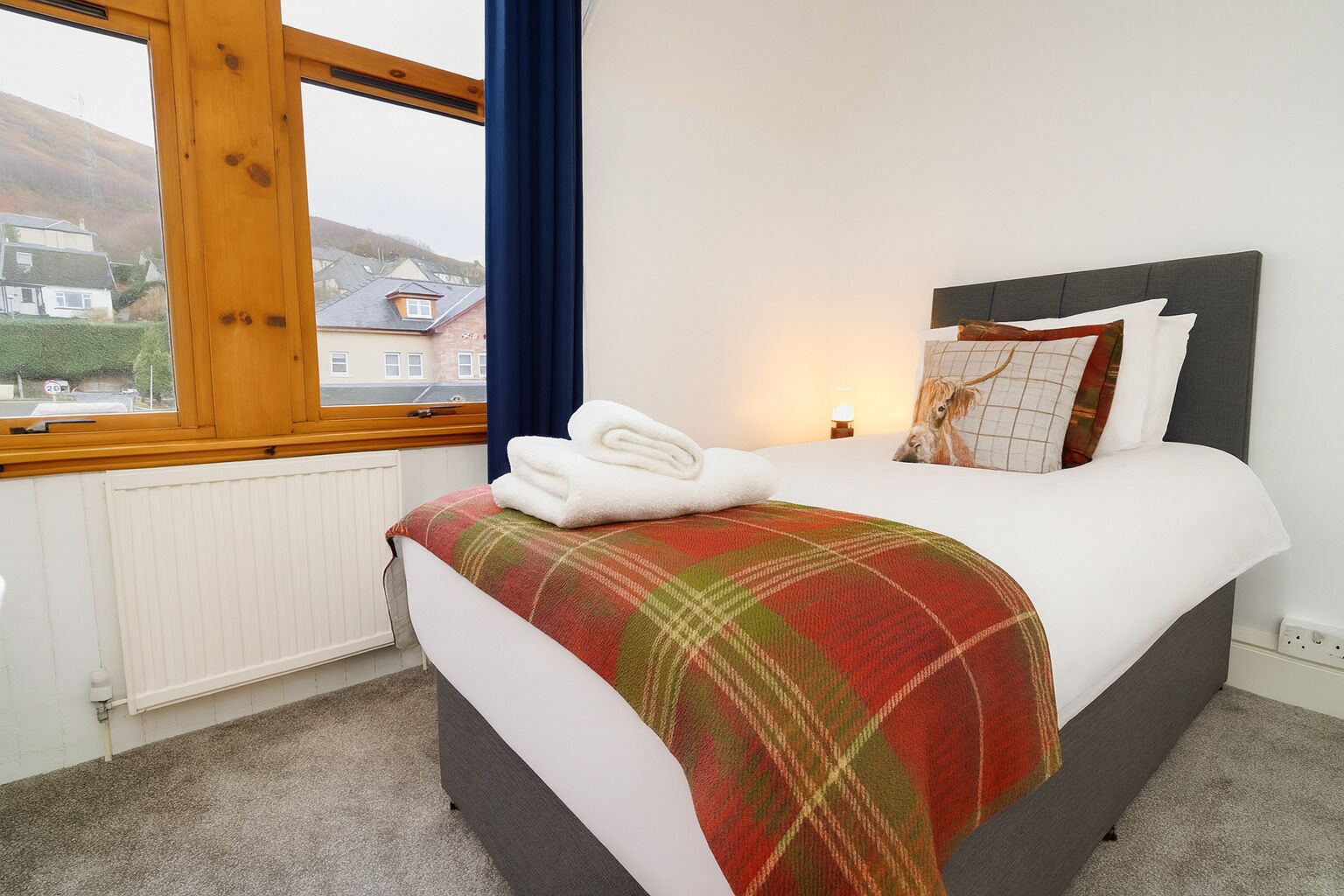 A bedroom with a bed, a window, a radiator, and a view of houses outside.