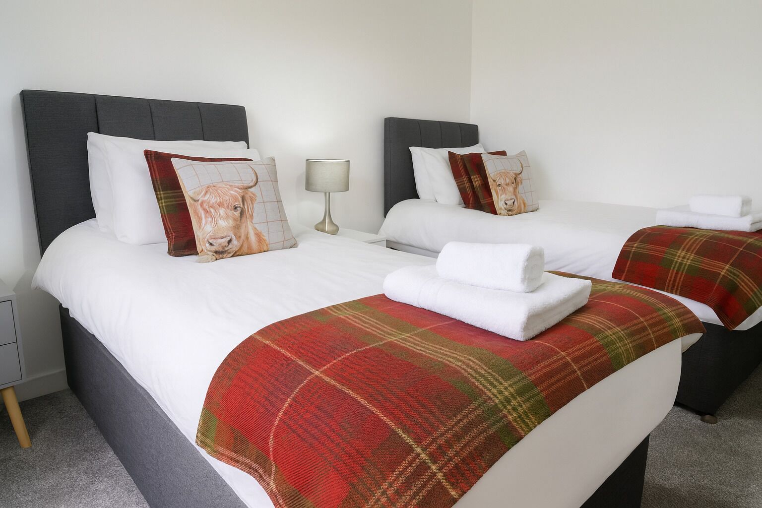 Habitación de hotel con dos camas, cada una con cabecero gris, ropa de cama blanca y una manta roja de cuadros. Hay una lámpara en una mesita de noche y una toalla sobre cada cama.