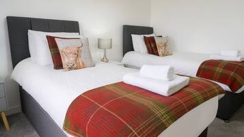 Habitación de hotel con dos camas, cada una con cabecero gris, ropa de cama blanca y una manta roja de cuadros. Hay una lámpara en una mesita de noche y una toalla sobre cada cama.