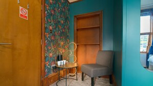 Ein Zimmer mit Türkiswänden, floralem Wandpapier, einer Holztür, einem grauen Sessel, einem kleinen runden Tisch mit Wasserkocher, Tassen und Teekanne sowie einem Rauchverbotsschild.
