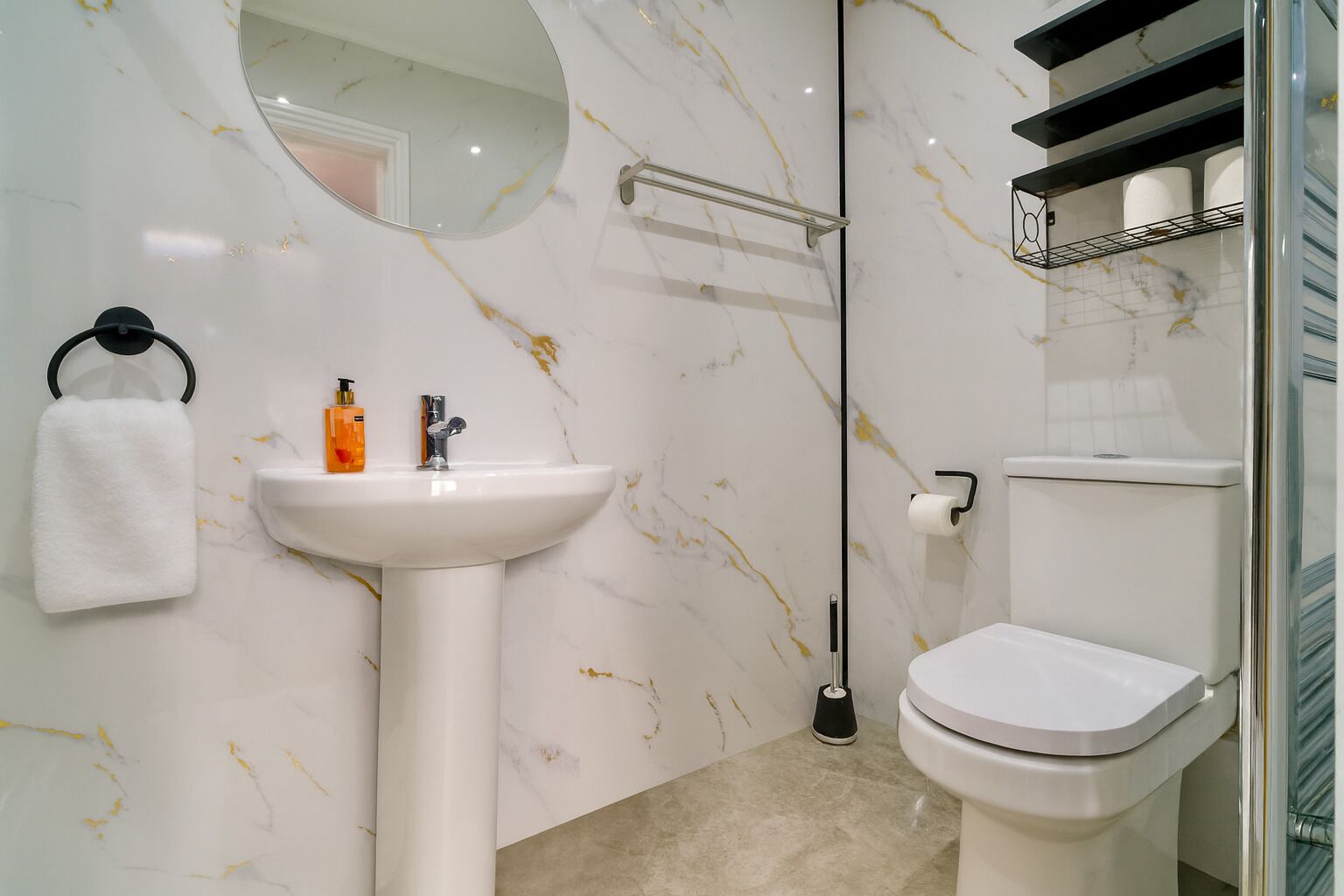 Un baño con pared de mármol, lavamanos pedestal, inodoro, toallero y estante con artículos de aseo.