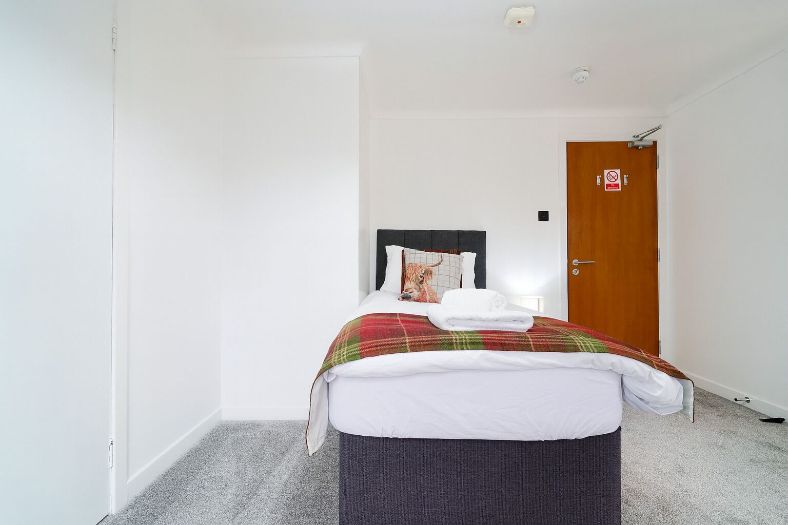 Una cama individual con una manta escocesa y un almohadón con la imagen de un perro, una puerta de madera con un cartel y una pared blanca.