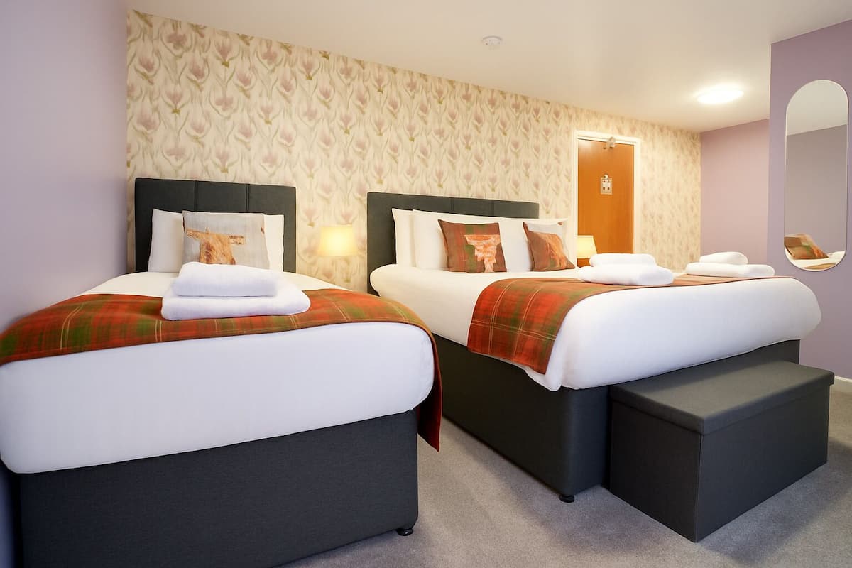 Habitación de hotel con dos camas, cada una con cabecero oscuro, ropa de cama blanca y manta a cuadros. Hay una mesita de noche con lámpara, un espejo en la pared y una puerta con interruptor de luz.