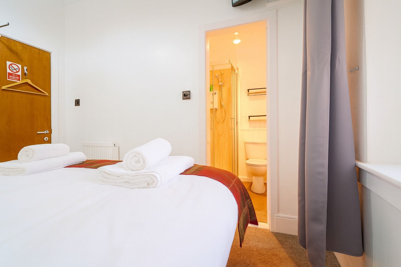 A hotel room with a bed, towels, a wooden door, a radiator, and a bathroom visible through an open door.