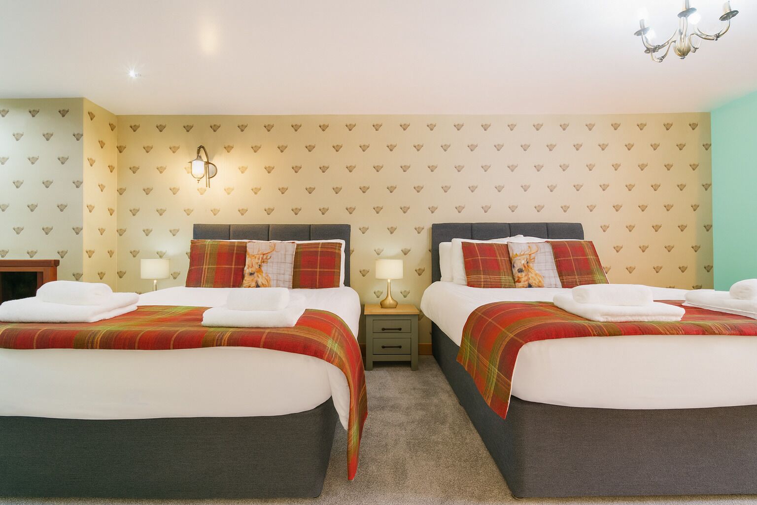 A hotel room with two beds, each with a plaid bedspread and matching pillows. There are bedside tables with lamps, a chandelier, and a patterned wallpaper.