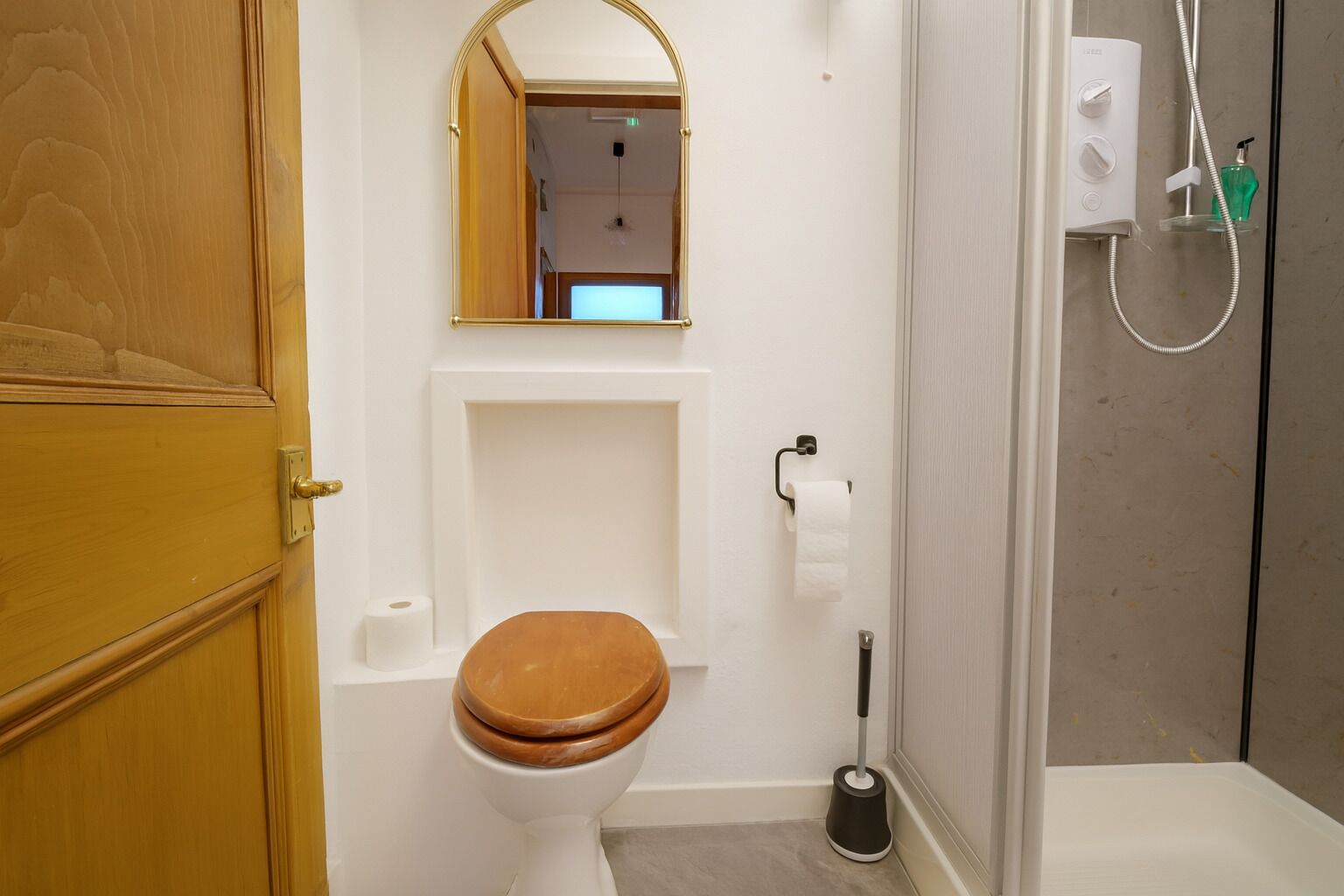 Un baño con puerta de madera, un inodoro con asiento de madera, un espejo, una zona de ducha con cabezal de ducha manual y un dispensador de papel higiénico.