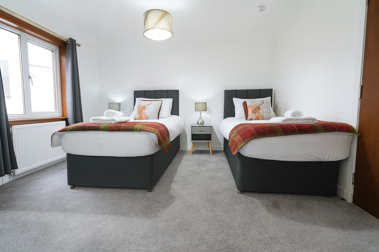 A hotel room with two beds, a small bedside table with a lamp, a radiator, and a window with curtains.