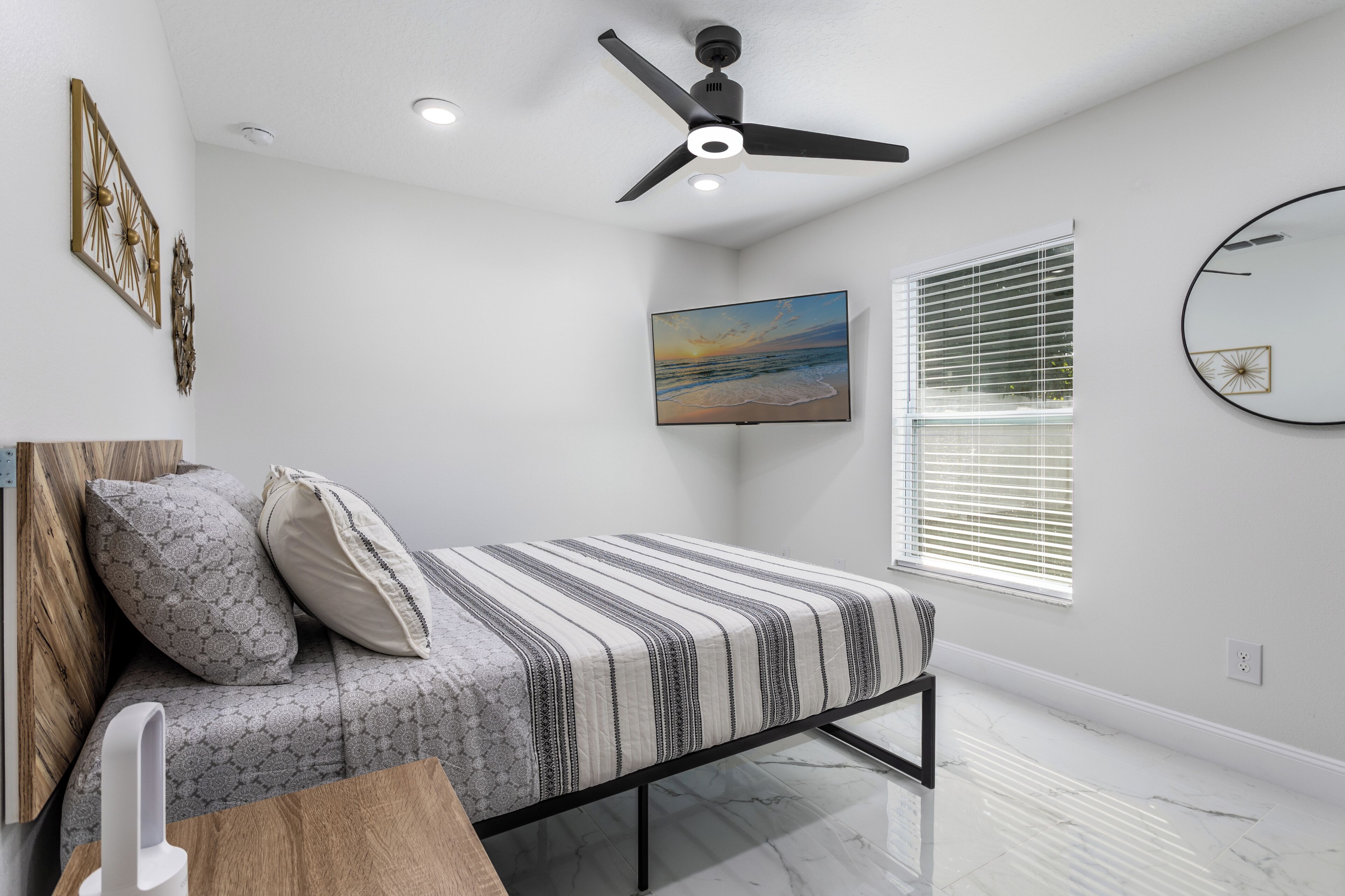 A bedroom with a bed, a ceiling fan, a painting, a window with blinds, and a wall-mounted mirror.
