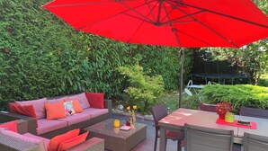Une terrasse extérieure avec des meubles en osier, un parasol rouge et une haie verdoyante en arrière-plan.