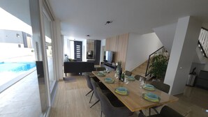 Un área de comedor moderna con una mesa larga dispuesta para seis personas, rodeada de sillas oscuras. Se observan al fondo una cocina y un salón, con una escalera que lleva a un nivel superior.