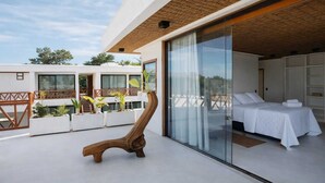 A modern hotel room with a large sliding glass door leading to an outdoor balcony with plants and a wooden chair. - Vila Maremangue  (Porto Seguro)
