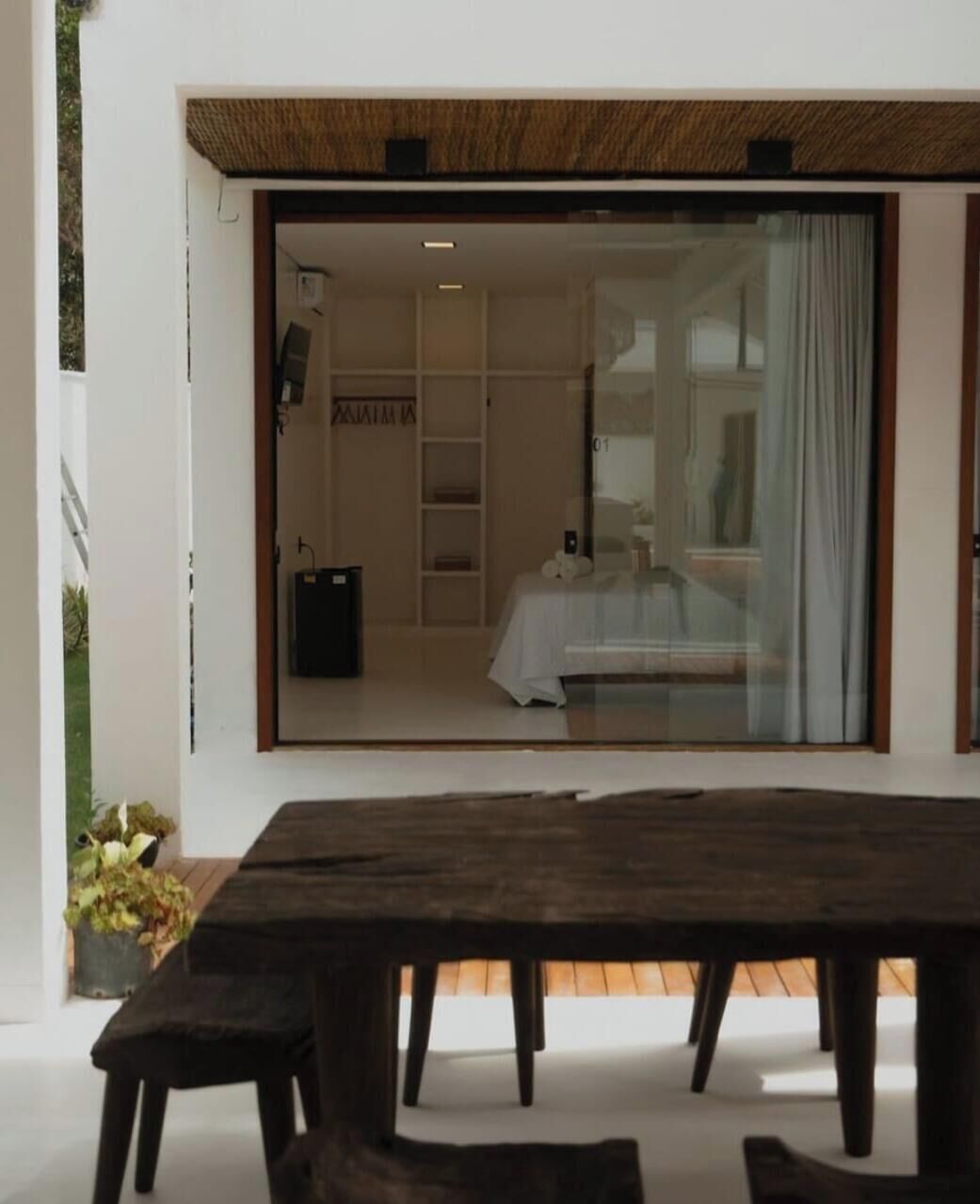 Un interior moderno con una mesa de comedor de madera, un ventanal de vidrio que da a un dormitorio y una planta pequeña a la izquierda.
