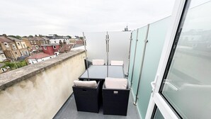 A balcony with a table and two wicker chairs, a view of the cityscape, and a glass door. - Stylish Penthouse in Willesden, Scrubs Lane (London)