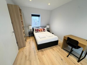 Een slaapkamer met een bed, een bureau en een stoel. Er is een raam met uitzicht op gebouwen.