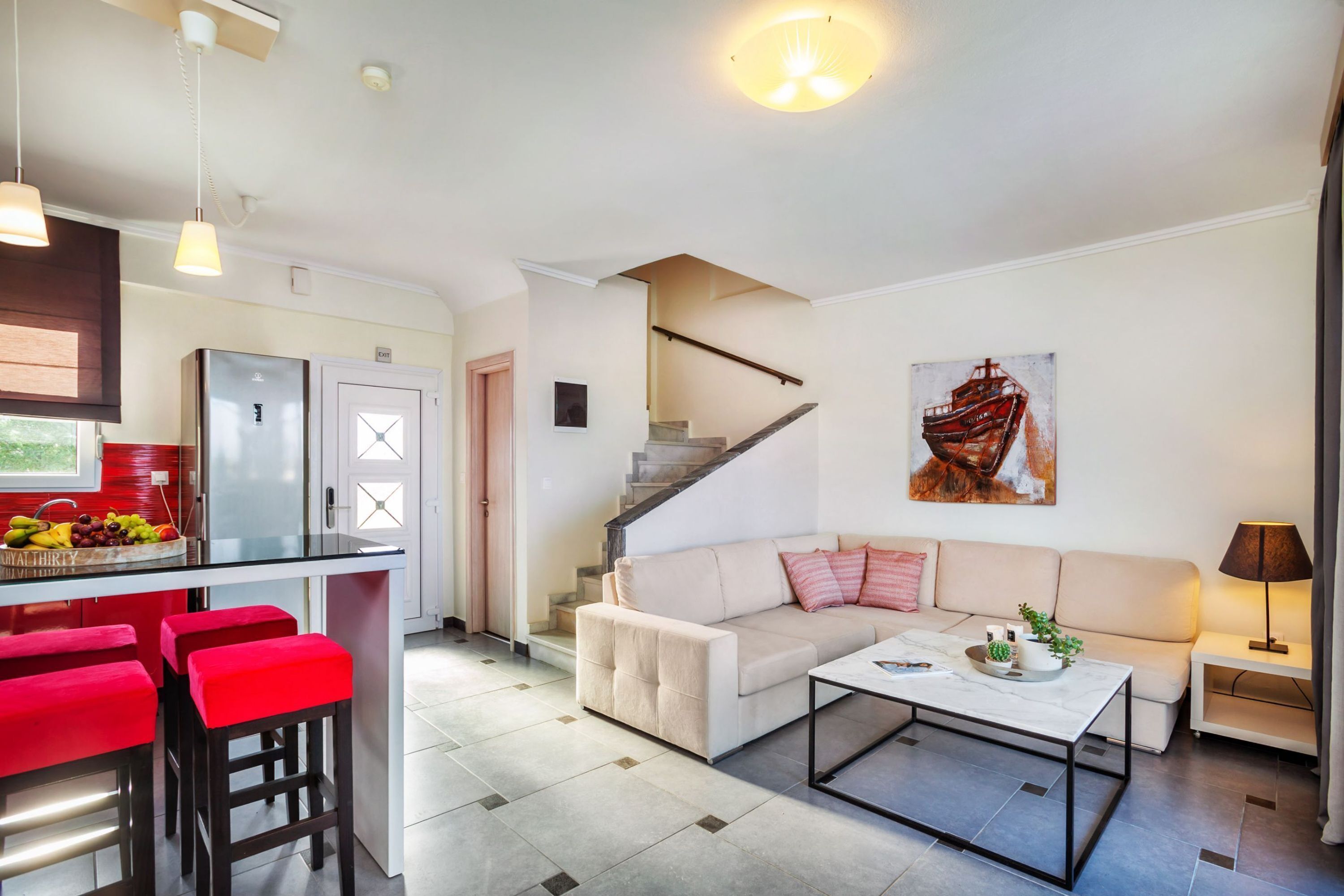 Et moderne køkken med et køleskab, et spiseområde med røde barstole, en stue med sofa og sofabord, og en trappe, der fører op til en etage.