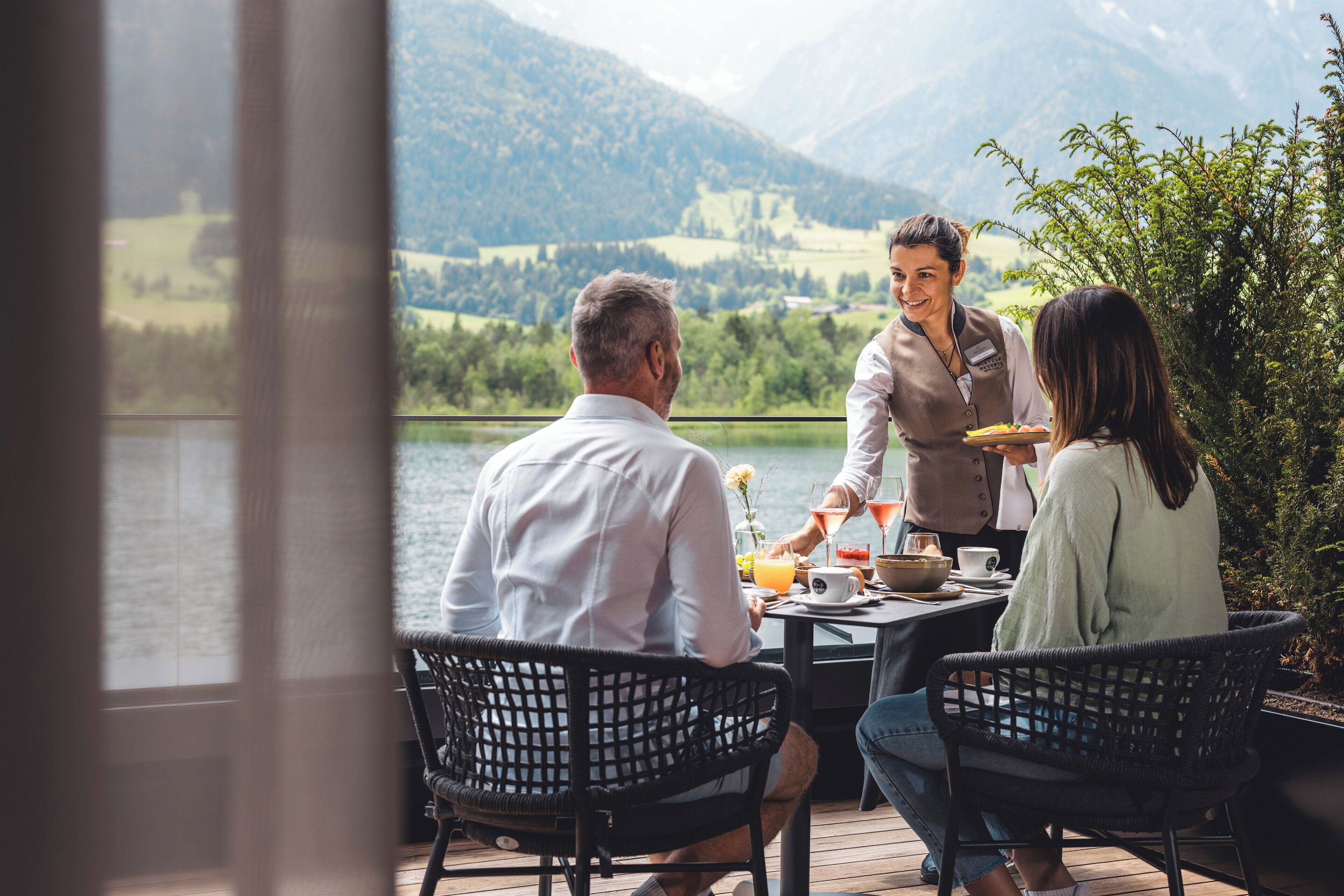 A dining area with a view of a lake and mountains, featuring a waiter serving food to guests seated at a table.
