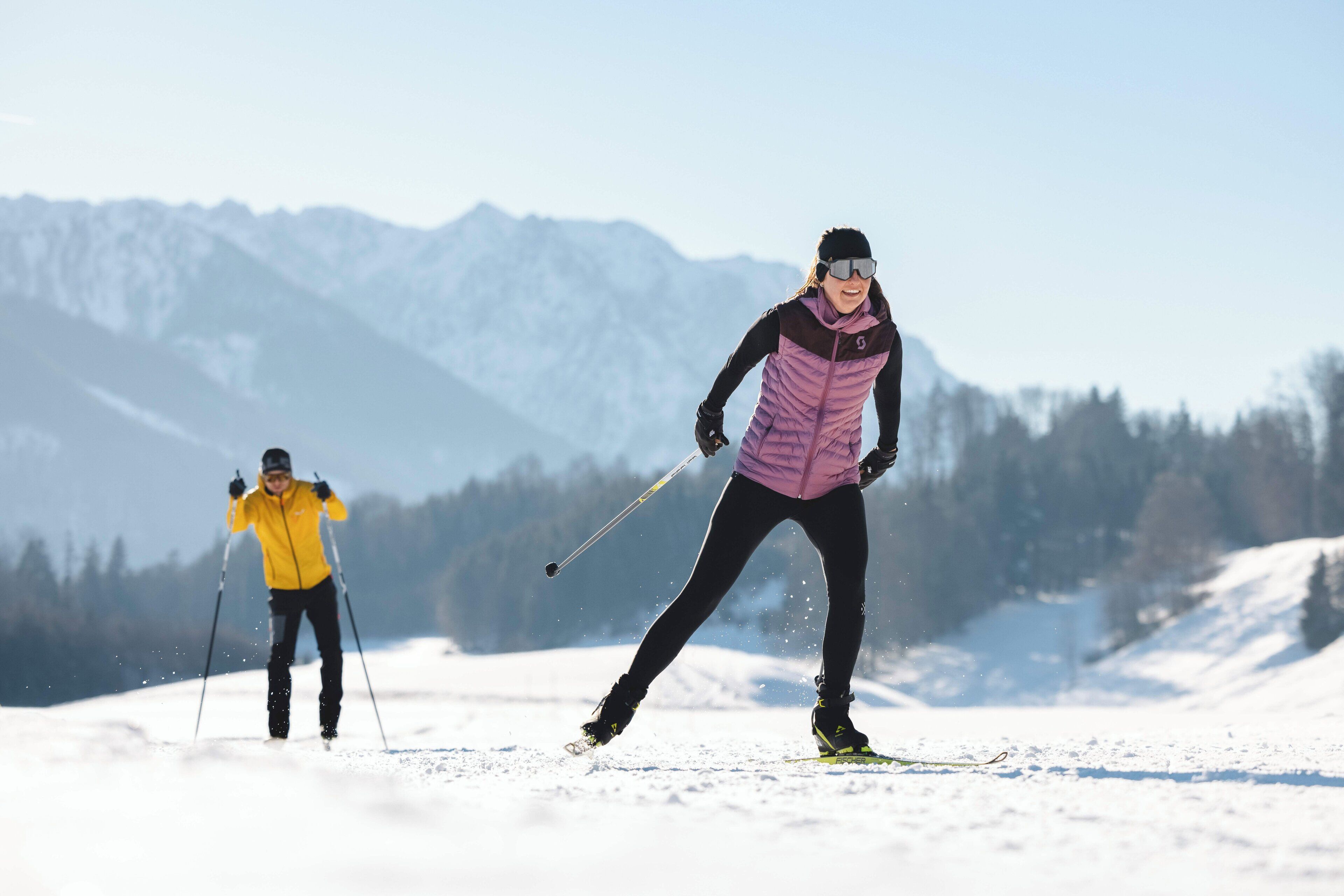 Two people skiing on a snowy mountain with a clear sky and distant mountains in the background.