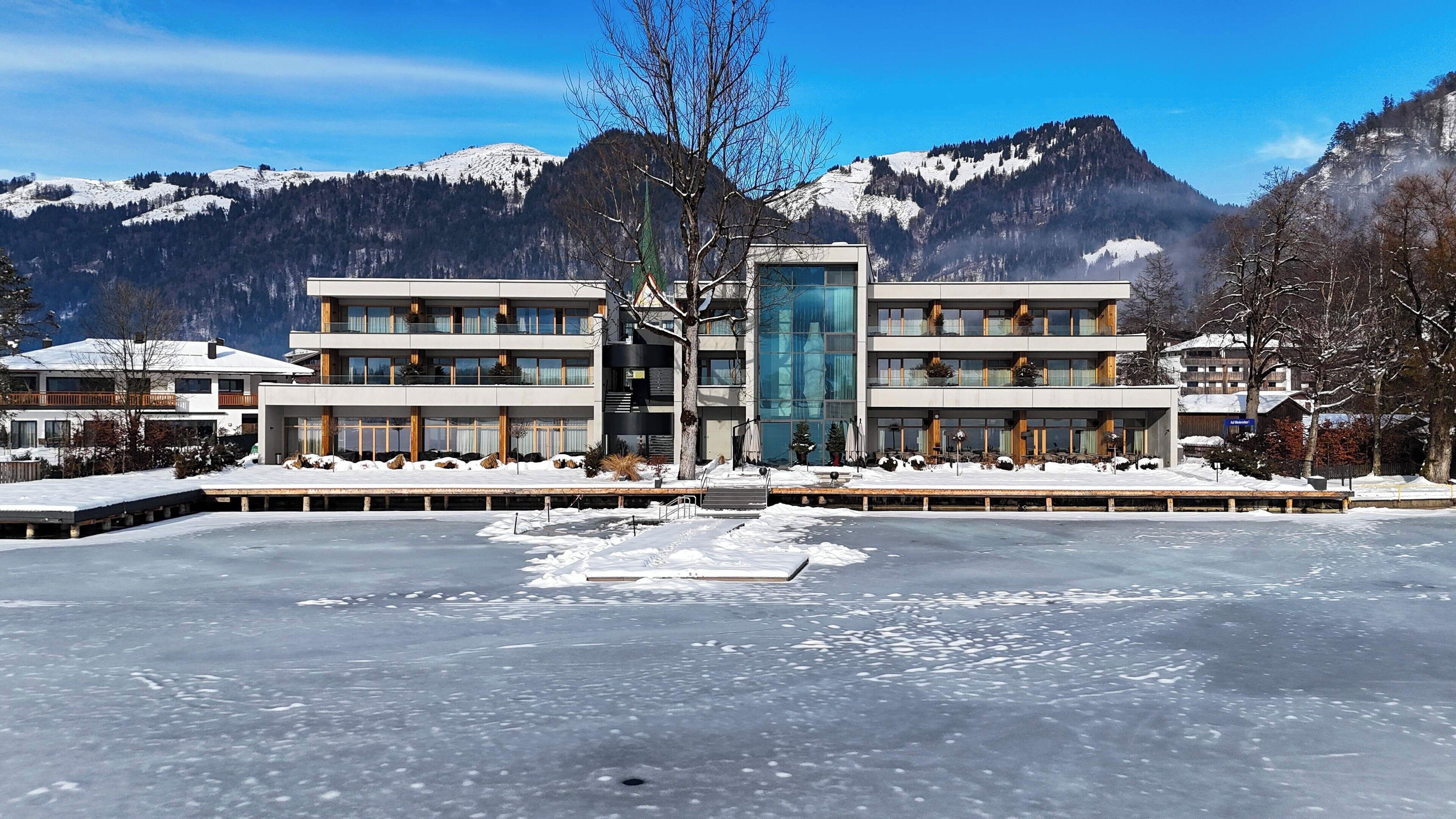 Ett modernt hotell med ett centralt placerat glastorn, flera balkonger och utsikt över snötäckta berg.