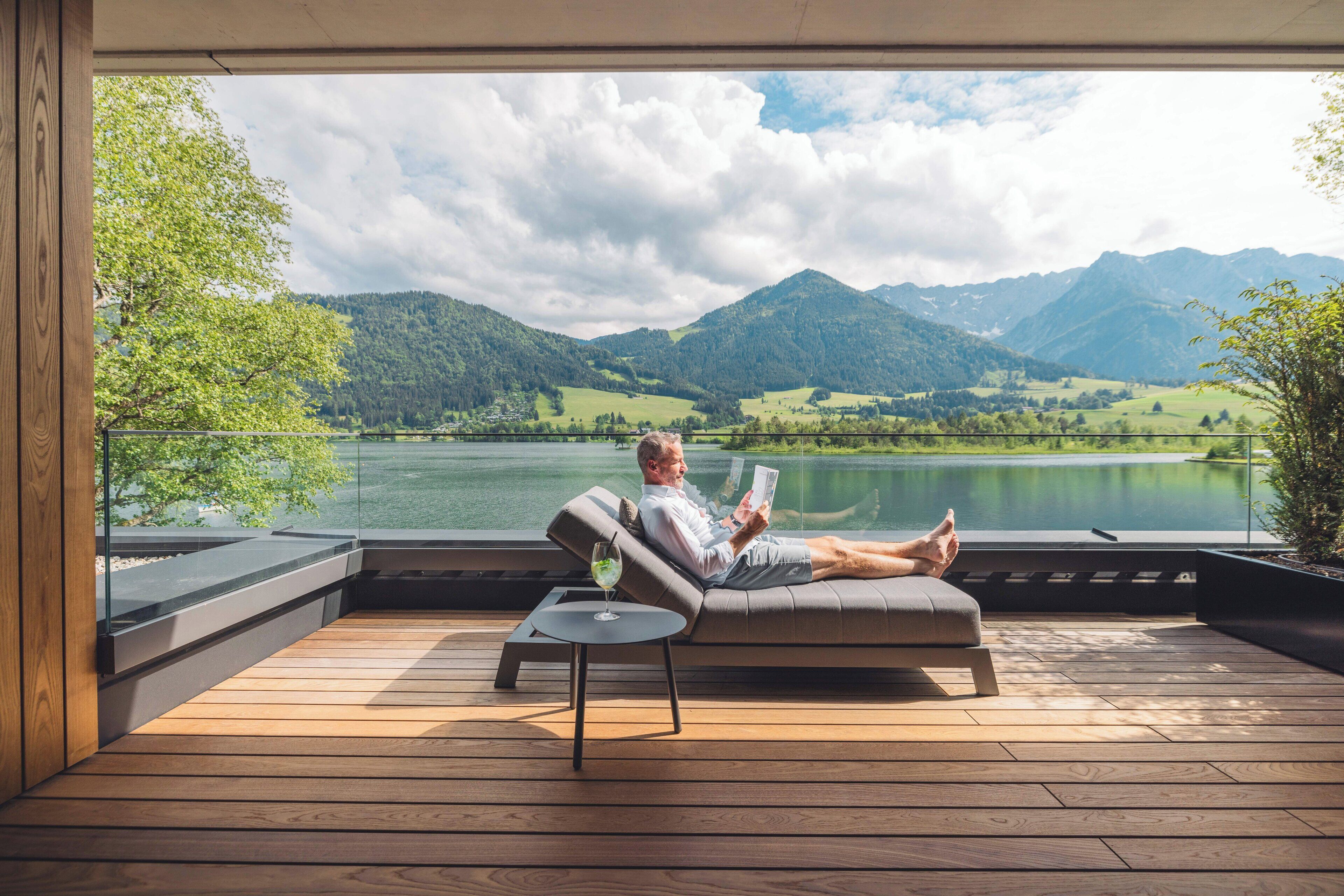 A man relaxing on a sofa with a book, overlooking a lake and mountains.