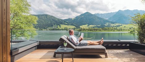 Un homme se détend sur un canapé, un livre à la main, avec vue sur un lac et des montagnes.