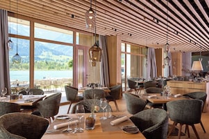 Un restaurante con vigas de madera en el techo, amplias ventanas que ofrecen vistas a montañas y agua, y mobiliario moderno.