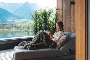 A person sitting on a sofa with a blanket, reading a book, with a view of mountains and a lake. - Lakeside Move&Relax (Walchsee)
