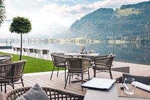 Un espace de restauration en plein air, avec des chaises en osier et une table dressée pour deux, offrant une vue sur un lac paisible et des collines boisées.