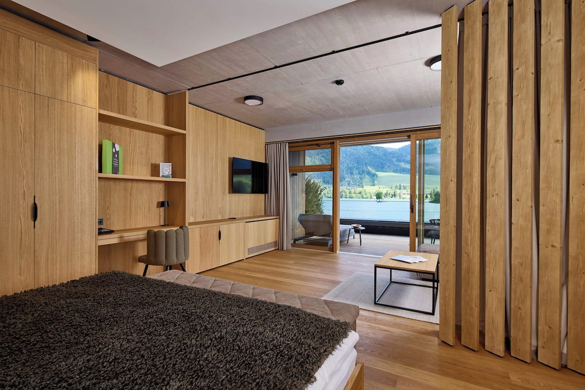 A modern bedroom with a large bed, wooden wardrobe, a desk, and a view of a lake through a sliding glass door.