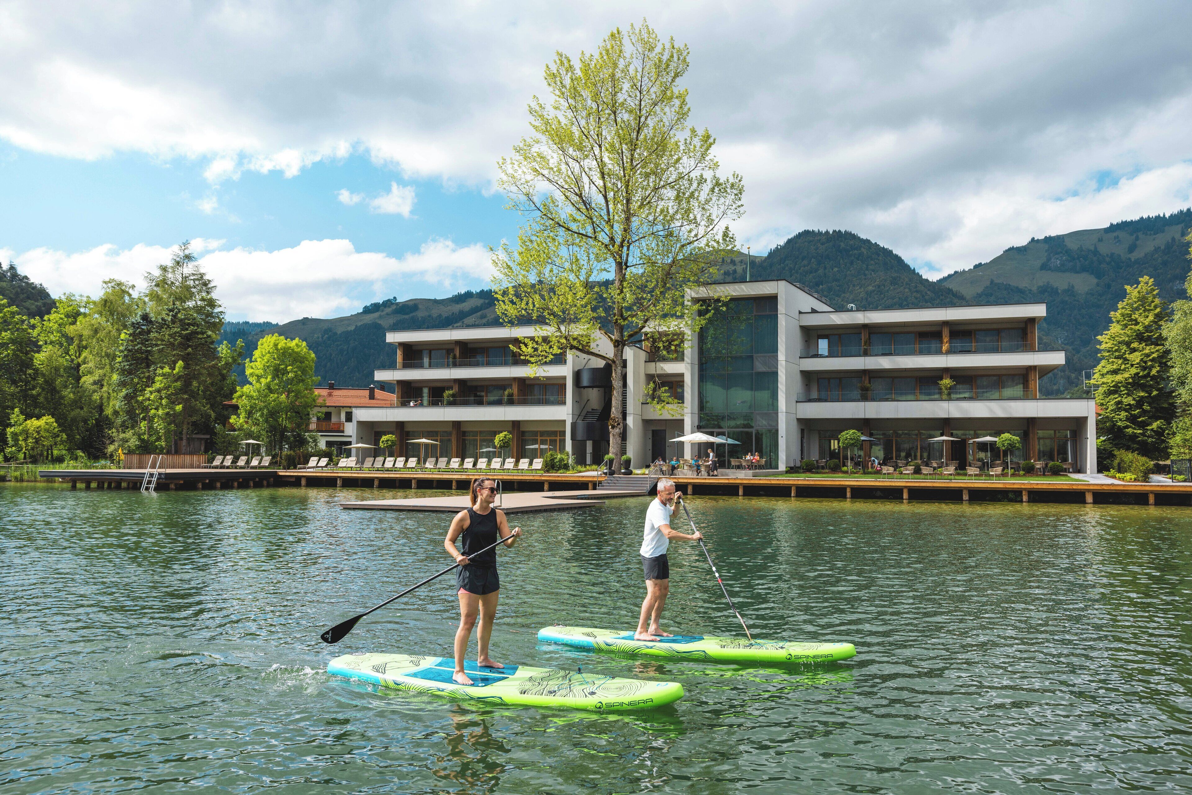 Two people paddleboarding on a lake with a modern building and mountains in the background.
