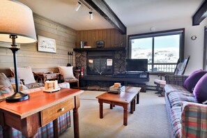 Ein gemütliches Wohnzimmer mit Kamin, Holzwänden, einer Couch, einem Couchtisch und Blick durch ein großes Fenster.