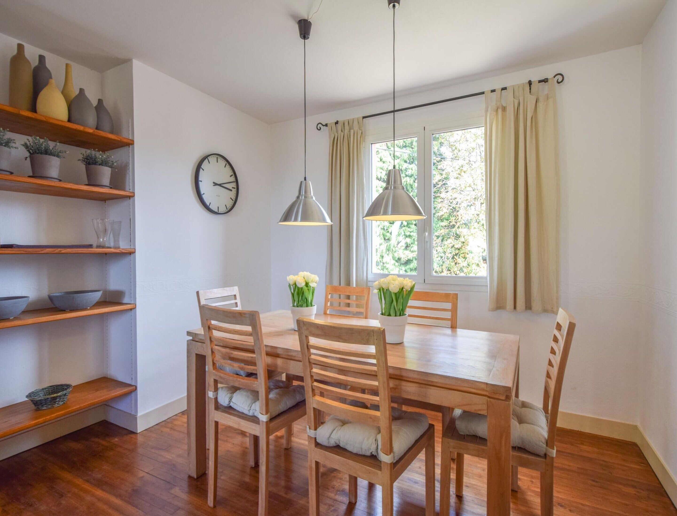 A dining area with a wooden table and chairs, a clock on the wall, and shelves with decorative items.